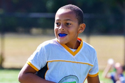 A boy playing a sport outdoors with a blue mouthguard in his mouth.